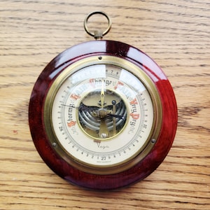 May include: A round analog barometer with a dark polished wood frame and a brass bezel. The cream-colored dial features the text Stormy, Change, Fair, and Very Dry, along with the brand name Vega and West Germany. It includes a metal hanging ring.