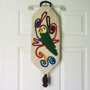 Puede incluir: Un tapiz decorativo para colgar en la pared con un loro verde con pico amarillo y detalles blancos, rodeado de espirales coloridas. El loro está bordado sobre una tela beige, con detalles de hilo rojo, azul y amarillo. Una borla marrón oscuro cuelga en la parte inferior.