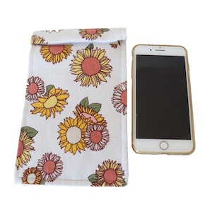 Op de afbeelding: Een witte telefoonhoes van stof met een zonnebloemmotief. De hoes heeft een klep die over de bovenkant van de telefoon vouwt.