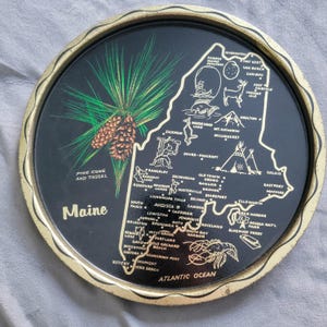 May include: A round black tray with a gold rim featuring a map of Maine. The tray has illustrations of a pine cone, lobster, and various Maine landmarks. The words "Maine" and "Atlantic Ocean" are also visible.