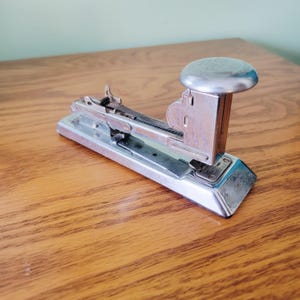 May include: A vintage silver stapler with a rounded top and the word "PILOT" embossed on it. The stapler has a rectangular base and a lever mechanism. The word "ACME" is visible on the base. The stapler is on a wooden surface.