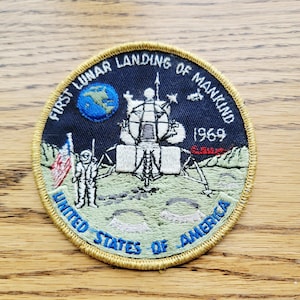 Peut inclure: Un écusson brodé circulaire sur fond bleu marine avec le texte FIRST LUNAR LANDING OF MANKIND et UNITED STATES OF AMERICA. Il représente un module lunaire blanc, un astronaute, un drapeau américain, la Terre et l'année 1969.