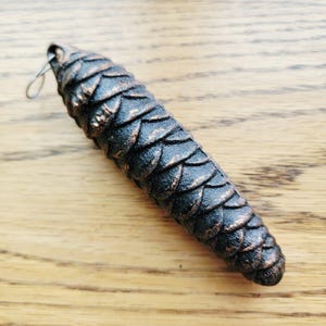 May include: A pendant shaped like a pinecone with a dark black finish and copper-colored highlights on the edges of the scales. The object features a small silver-toned metal jump ring attached to the top for hanging or connecting to a chain.