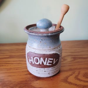 May include: A ceramic honey pot with a blue lid and a wooden honey dipper. The pot is off-white with a brown band that reads "HONEY". The rim and lid are blue. The honey dipper is light brown.