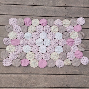 May include: A handmade quilt featuring a pattern of connected circles. The circles are in shades of pink, white, and floral prints, arranged on a wooden surface. The quilt's design is symmetrical, creating a visually appealing textile.