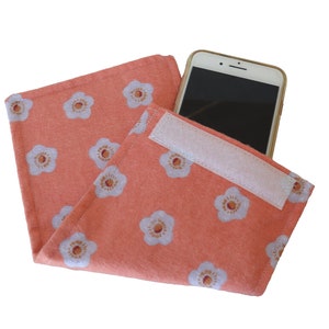 Op de afbeelding: Een perzikkleurige stoffen tas met een wit bloemenpatroon en een witte klittenbandsluiting. Een witte smartphone zit in de tas.