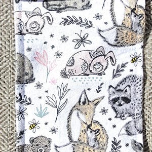 Op de afbeelding: Witte stoffen etui met een herhalend patroon van bosdieren, waaronder vossen, konijnen, wasberen en beren. Het ontwerp bevat bloemen- en bladeren in zwart, roze en groen, waardoor een grillige, door de natuur geïnspireerde esthetiek ontstaat.