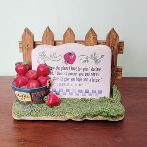 May include: A decorative figurine featuring a wooden fence, a sign with a Bible verse, and a barrel of red apples. The sign has a red apple illustration and the text "how the plans I have for you." The barrel reads "Apples 5¢."