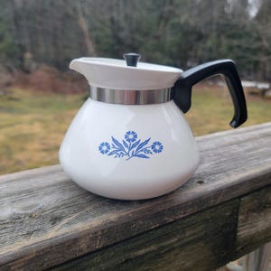May include: White ceramic coffee pot with a black handle and lid. The pot features a blue floral design and a silver band around the middle. The pot is sitting on a wooden surface.
