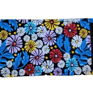 Op de afbeelding: Portemonnee met een bloemenpatroon op een zwarte achtergrond. Het ontwerp bevat rode, gele, paarse en witte bloemen, met blauwe bladeren. Het heeft een zwarte klittenbandsluiting. Ongeveer 15 cm lang.