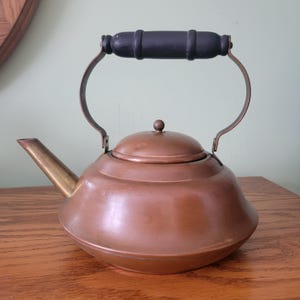 May include: A copper-colored kettle with a black wooden handle and a brass spout. The kettle has a rounded body and a lid with a small knob. The handle is attached to the kettle with curved metal supports. The kettle is sitting on a wooden surface.