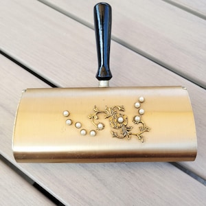 May include: A vintage gold-toned dustpan with a black handle. The dustpan is decorated with an ornate floral design and faux pearls. The handle is black and glossy. A decorative item for the home.