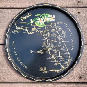 May include: A dark blue, round serving tray with a gold rim and a map of Florida. The map includes city names and illustrations of landmarks. The tray is decorated with orange blossoms and the words "Florida", "Gulf of Mexico", and "Atlantic Ocean".