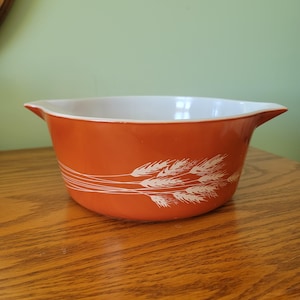 May include: An orange Pyrex mixing bowl with a white interior and a wheat stalk design. The bowl has a handle on each side and is set on a wooden surface. The wheat design is white.