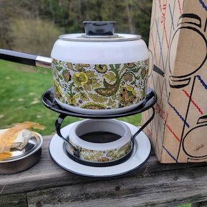 Könnte beinhalten: Vintage Fondue-Topf aus weißem Emaille mit schwarzem Griff und Deckel. Der Topf hat ein farbenfrohes Paisley-Design in Grün, Gelb und Braun. Er steht auf einem schwarzen Metallständer mit passendem Wärmer. Ein Retro-Küchenartikel.