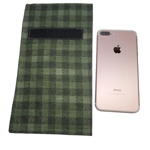 Op de afbeelding: Een groen en zwart geruite telefoonhoes met een zwarte klittenbandsluiting ligt naast een roségouden iPhone. De hoes is rechthoekig en lijkt van stof te zijn. De iPhone heeft een zilveren camera en het Apple-logo.