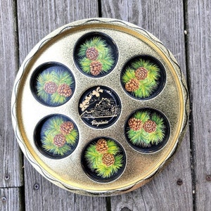 May include: Gold-colored metal tray with six round compartments, each featuring a black and white illustration of pine cones. The tray has a raised rim and a central circular area with the word "Virginia" in gold lettering.