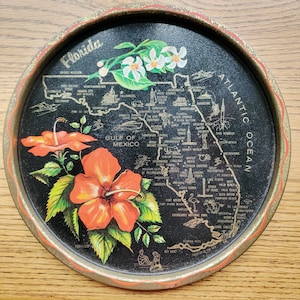 May include: Round vintage Florida souvenir tray with a black background. The tray features a map of Florida with gold lettering and illustrations of landmarks. It is decorated with orange hibiscus flowers and white blossoms. The tray has a gold rim.