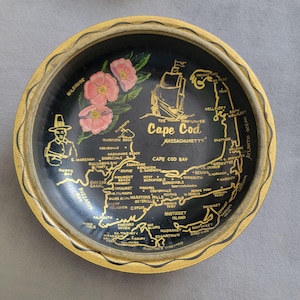 May include: A round, black decorative plate with a gold rim featuring a map of Cape Cod, Massachusetts. The plate includes illustrations of a ship, flowers, and a pilgrim. The text includes the words "Cape Cod" and various town names.