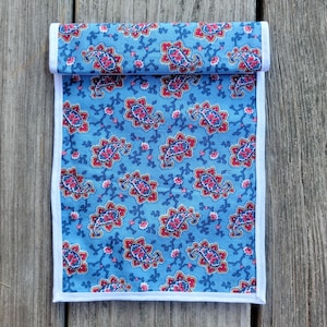 Op de afbeelding: Een blauwe stoffen loper met een paisley-patroon in rood, wit en bruin. De loper heeft een witte rand.