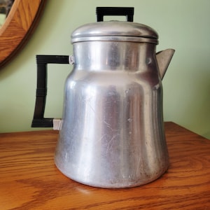 Puede incluir: Cafetera vintage de aluminio con asa y asa de tapa negras. La cafetera tiene un pico y un cuerpo redondeado, sobre una superficie de madera. La cafetera mide aproximadamente 25 cm de altura.