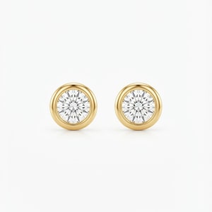 May include: A pair of stud earrings featuring round, clear, faceted stones set in polished yellow gold bezels. Each earring has a smooth, circular gold frame surrounding the central stone, creating a clean, classic look with a reflective finish.