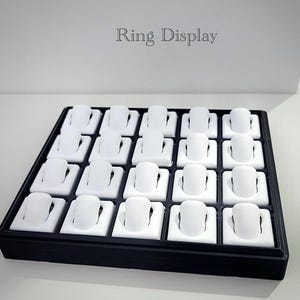 Può includere: Un espositore per anelli nero con 20 supporti bianchi. Il testo "Ring Display" è stampato in grigio in alto. Il vassoio è rettangolare e ha una cornice nera.