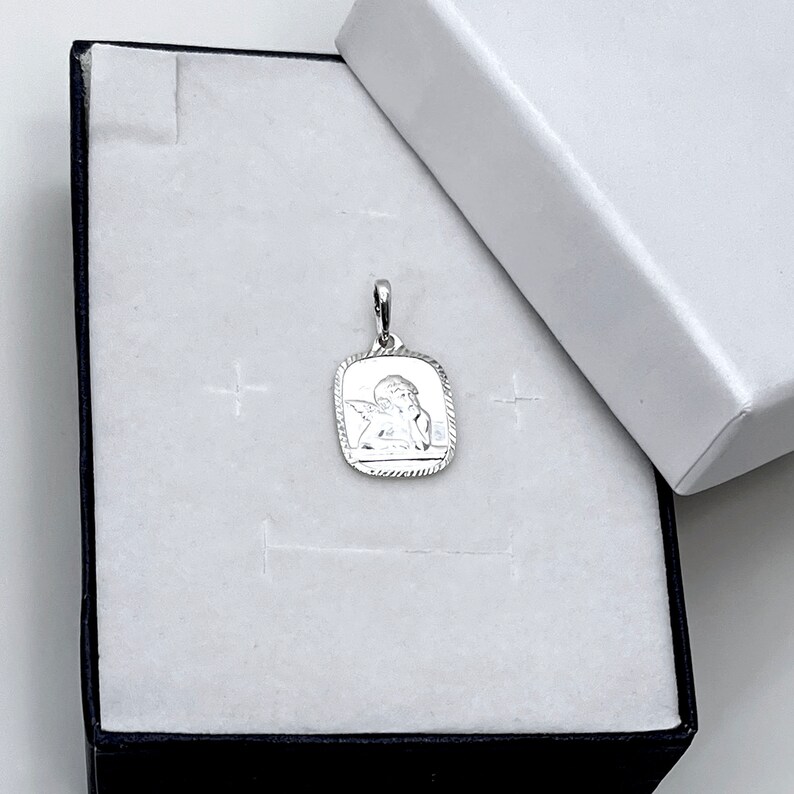 May include: A silver pendant featuring a detailed image of a cherub angel with wings. The pendant is in a white box with a black border.