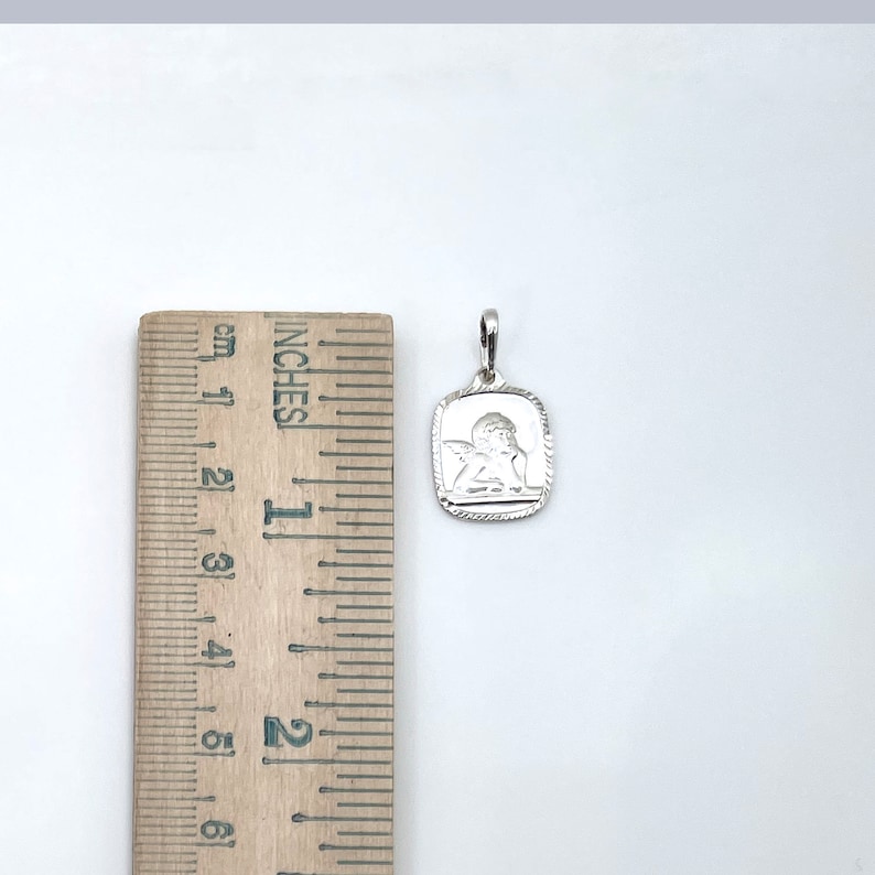 May include: Silver pendant charm featuring a detailed angel design. The pendant is square shaped with a textured border and measures approximately 1 inch in length.