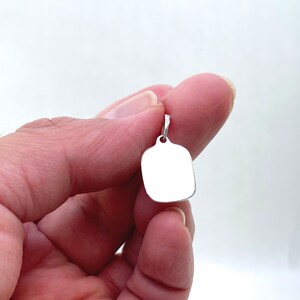 May include: A silver blank tag pendant with a rounded rectangular shape. The pendant has a loop at the top for attaching to a chain.