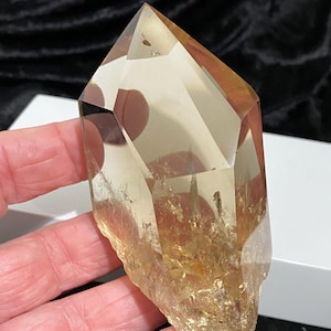 Op de afbeelding: Een grote, gefacetteerde, goudgele kwartskristal met een punt. Het kristal heeft een ruwe, natuurlijke basis en wordt in een hand gehouden. De kleur van het kristal varieert van lichtgeel tot een diepere amberkleur, met zichtbare insluitsels.