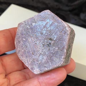 Pode incluir: Uma gema bruta, não cortada, com uma forma geométrica multifacetada. A pedra tem um tom roxo claro com variações sutis de cor e textura. A superfície exibe padrões intrincados e uma aparência ligeiramente cristalina.