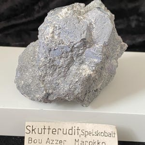 Può includere: Un campione di Skutterudite grigio ruvido con lucentezza metallica e sfumature di blu e viola. Il campione minerale è posto su una superficie bianca, con un'etichetta che recita "Skutterudit,speiskobalt Bou Azzer Marokko."