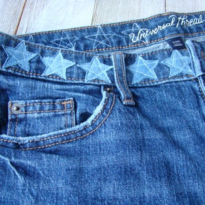 May include: Blue denim jeans with a waistband featuring a row of blue star-shaped embellishments. The waistband also has the text "Universal Thread" embroidered in white.