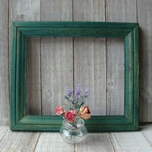May include: A teal green wooden picture frame with a simple design. The frame is sitting on a light wood surface with a small glass vase filled with dried flowers.