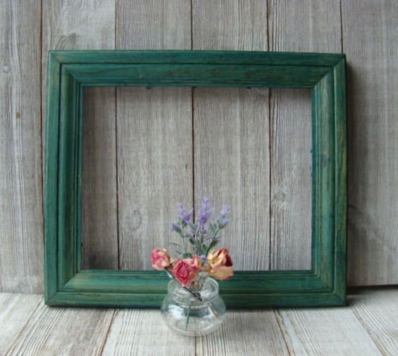 May include: A teal green wooden picture frame with a simple design. The frame is sitting on a light wood surface with a small glass vase filled with dried flowers.