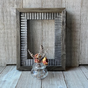 May include: A rustic, rectangular picture frame with a corrugated metal insert. The frame is made of weathered wood. A small glass jar with dried flowers sits in front of the frame, adding a touch of natural decor.