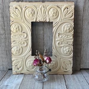 May include: A rectangular cream-colored metal frame featuring an embossed floral and scroll pattern with distressed edges. A small clear glass jar with a wire handle sits in front, containing dried pink roses and sprigs of lavender.