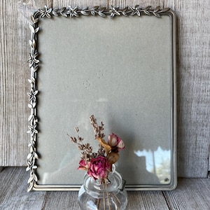 May include: A rectangular silver-toned metal picture frame featuring a decorative border of embossed flowers and leaves along the top and left edges. In front of the frame sits a small, clear glass jar containing a bouquet of dried pink roses.