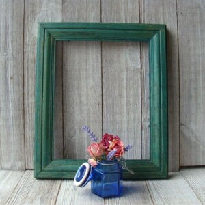 May include: A blue glass jar with a lid and a floral arrangement of dried flowers inside. The jar is sitting in front of a green picture frame.