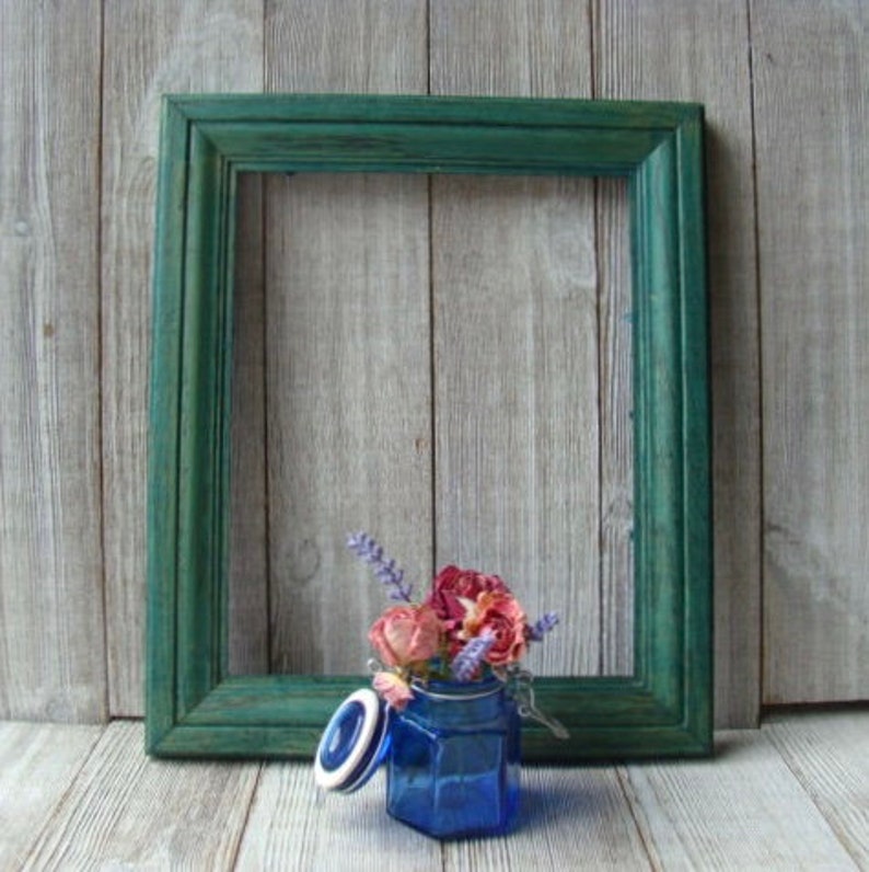 May include: A blue glass jar with a lid and a floral arrangement of dried flowers inside. The jar is sitting in front of a green picture frame.