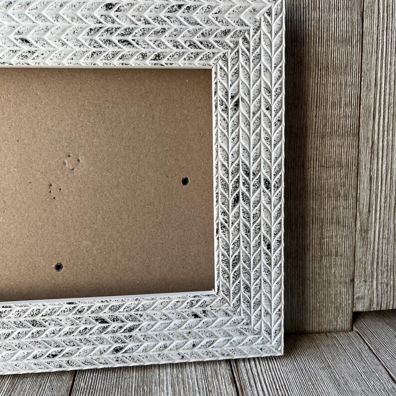 May include: A white rectangular picture frame with a distressed finish. The frame has a detailed leaf pattern around the edges. The interior of the frame is a light brown color, and the frame is set against a wooden background.