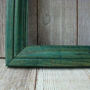 May include: A close-up of a dark green wooden picture frame with a visible grain pattern. The frame is against a light wood background.