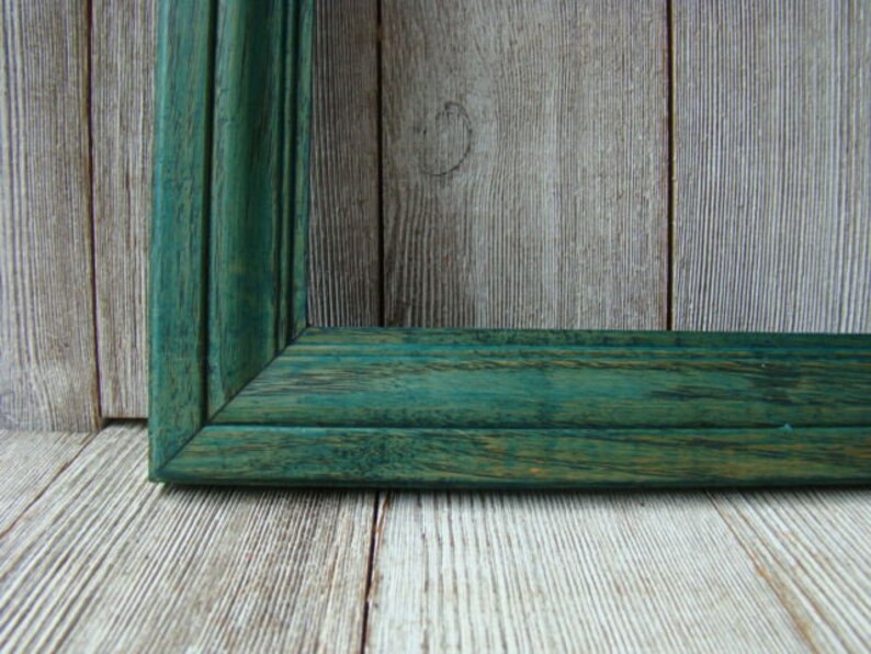 May include: A close-up of a dark green wooden picture frame with a visible grain pattern. The frame is against a light wood background.