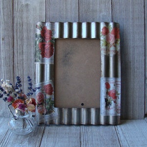 May include: A silver corrugated metal picture frame with floral accents and a small glass vase with dried flowers. The frame has a rectangular opening and is decorated with red roses and vintage-style labels. The vase contains lavender and other dried flowers.