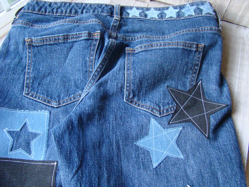 May include: Blue denim jeans with a row of blue stars across the waistband and several star-shaped patches sewn on the back pockets. The patches are in various shades of blue and black.