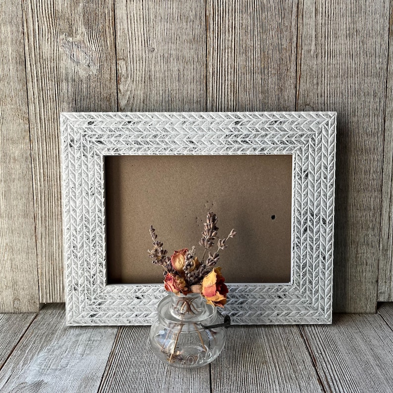 May include: A white rectangular picture frame with a textured, chevron-like pattern. A small glass vase holds dried flowers in front of the frame. The background is a wooden surface.