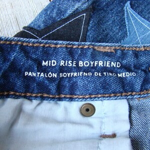 May include: Close-up of a blue denim jean waistband with a white label that reads "Mid Rise Boyfriend Pantal&oacute;n Boyfriend de Tiro Medio".