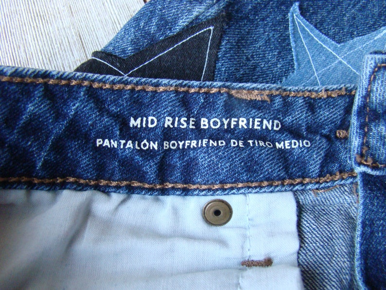 May include: Close-up of a blue denim jean waistband with a white label that reads "Mid Rise Boyfriend Pantal&oacute;n Boyfriend de Tiro Medio".