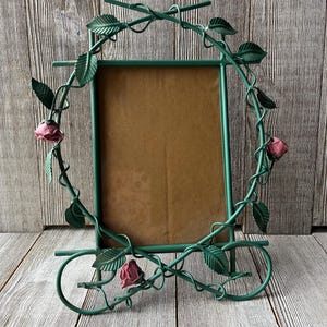 May include: A green metal picture frame with a floral design. The frame features a rectangular opening with a brown backing. The frame is adorned with green leaves and pink roses, creating a decorative and elegant look.